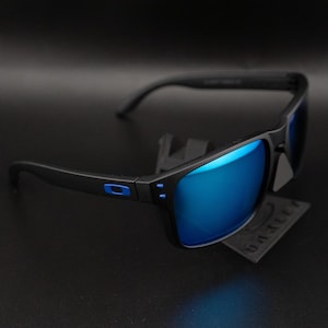 May include: Black sunglasses with blue mirrored lenses. The frames are black with a blue accent on the temple. The sunglasses are displayed on a black surface.