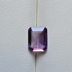May include: A rectangular, faceted amethyst gemstone with a deep purple hue. The gem is set against a textured, off-white background. The cut of the stone allows light to reflect, creating a sparkling effect.