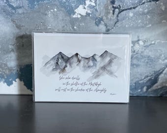May include: A greeting card featuring a watercolor painting of gray and black mountain peaks. The card includes the text "She who dwells in the shelter of the Most High will rest in the shadow of the Almighty."