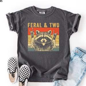 May include: Dark gray t-shirt with a raccoon graphic. The raccoon is giving the rock and roll hand gesture. The text "FERAL & TWO" is above the raccoon, with "EST. 2024" below. The shirt is paired with blue jeans and checkered sneakers.