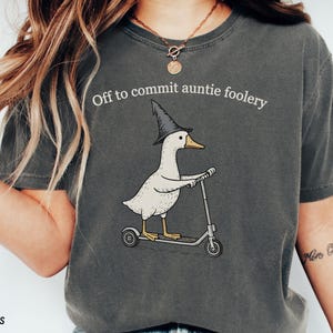 May include: Dark grey t-shirt with the phrase "Off to commit auntie foolery." The shirt features a cartoon goose wearing a witch hat and riding a scooter. The words "Comfort Colors Pepper" are printed at the bottom.