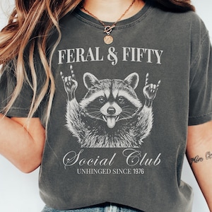 May include: A charcoal gray t-shirt featuring a white graphic of a raccoon making rock-and-roll hand gestures. The text reads FERAL & FIFTY, Social Club, and UNHINGED SINCE 1976. The shirt has a crew neckline and short sleeves in a vintage wash.