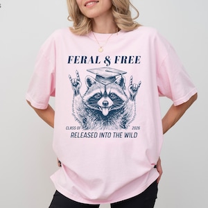 May include: Light pink t-shirt with a raccoon graphic wearing a graduation cap and the text "FERAL & FREE". The raccoon is making a rock and roll hand gesture. Below the raccoon is the text "CLASS OF 2026 RELEASED INTO THE WILD".