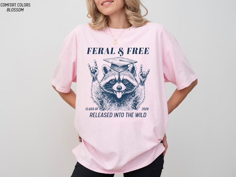 May include: Light pink t-shirt with a raccoon graphic wearing a graduation cap and the text "FERAL & FREE". The raccoon is making a rock and roll hand gesture. Below the raccoon is the text "CLASS OF 2026 RELEASED INTO THE WILD".