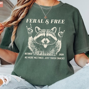 May include: Moss green t-shirt with a raccoon graphic. The raccoon is giving the rock on hand gesture. Text on the shirt reads "FERAL & FREE", "RETIRED 2026", and "NO MORE MEETINGS. JUST TRASH SNACKS".