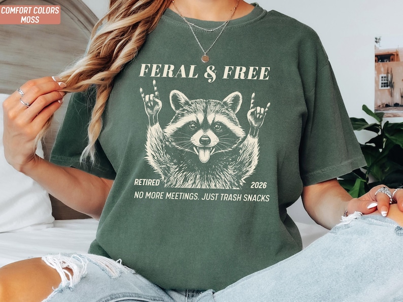 May include: Moss green t-shirt with a raccoon graphic. The raccoon is giving the rock on hand gesture. Text on the shirt reads "FERAL & FREE", "RETIRED 2026", and "NO MORE MEETINGS. JUST TRASH SNACKS".