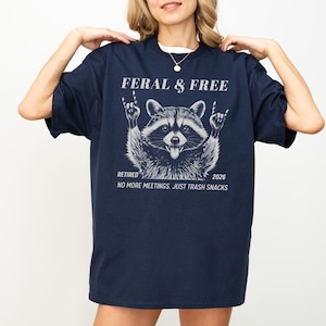 May include: Navy blue t-shirt with a raccoon graphic. The raccoon is making a rock and roll hand gesture. Text on the shirt reads "FERAL & FREE", "RETIRED 2026", and "NO MORE MEETINGS. JUST TRASH SNACKS".