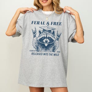 May include: A light gray oversized t-shirt with a navy blue graphic of a raccoon wearing a graduation cap and making a rock-and-roll hand gesture. The text reads "FERAL & FREE" and "CLASS OF 2026 RELEASED INTO THE WILD."