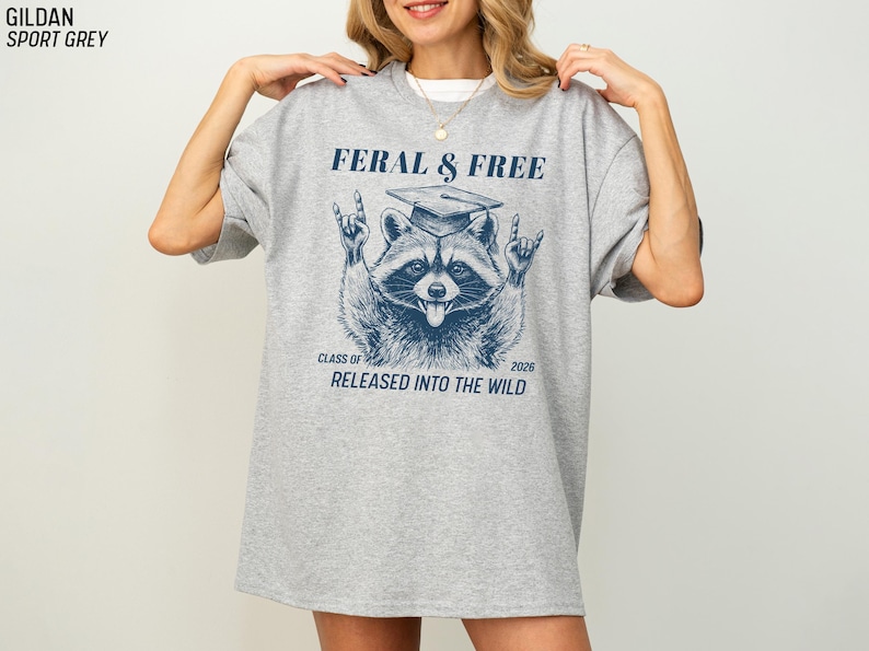 May include: A light gray oversized t-shirt with a navy blue graphic of a raccoon wearing a graduation cap and making a rock-and-roll hand gesture. The text reads "FERAL & FREE" and "CLASS OF 2026 RELEASED INTO THE WILD."