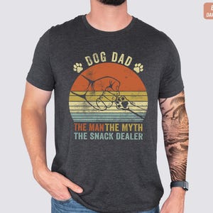 May include: Dark grey Bella Canvas t-shirt with a retro graphic. The design features the text "DOG DAD" above a paw print and a graphic of a hand shaking a dog's paw inside a sunset circle. Below the circle is the text "THE MAN THE MYTH THE SNACK DEALER".