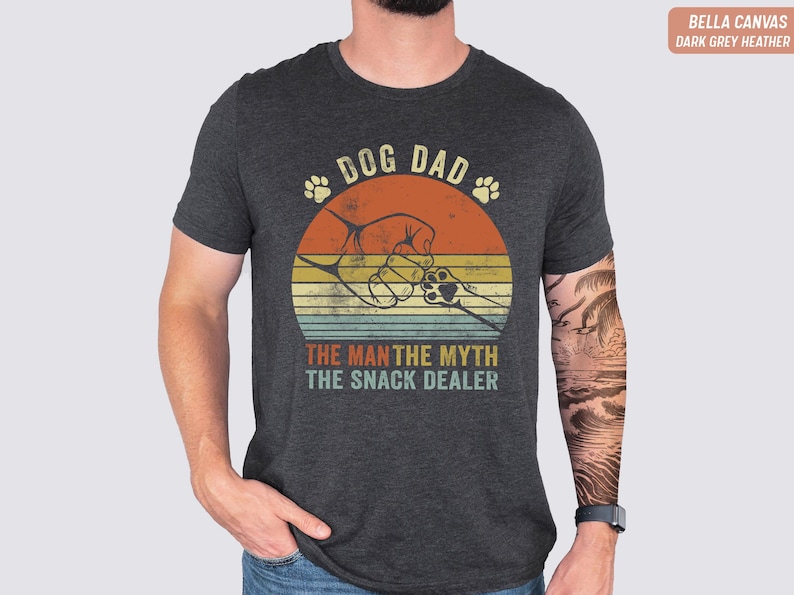 May include: Dark grey Bella Canvas t-shirt with a retro graphic. The design features the text "DOG DAD" above a paw print and a graphic of a hand shaking a dog's paw inside a sunset circle. Below the circle is the text "THE MAN THE MYTH THE SNACK DEALER".