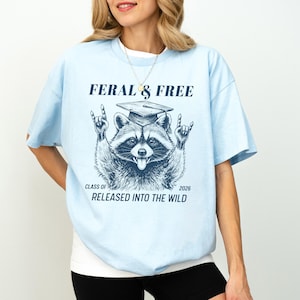 May include: Light blue t-shirt with a raccoon graphic wearing a graduation cap and the text "FERAL & FREE". The raccoon is making a rock and roll hand gesture. The text "CLASS OF 2026 RELEASED INTO THE WILD" is also printed on the shirt.