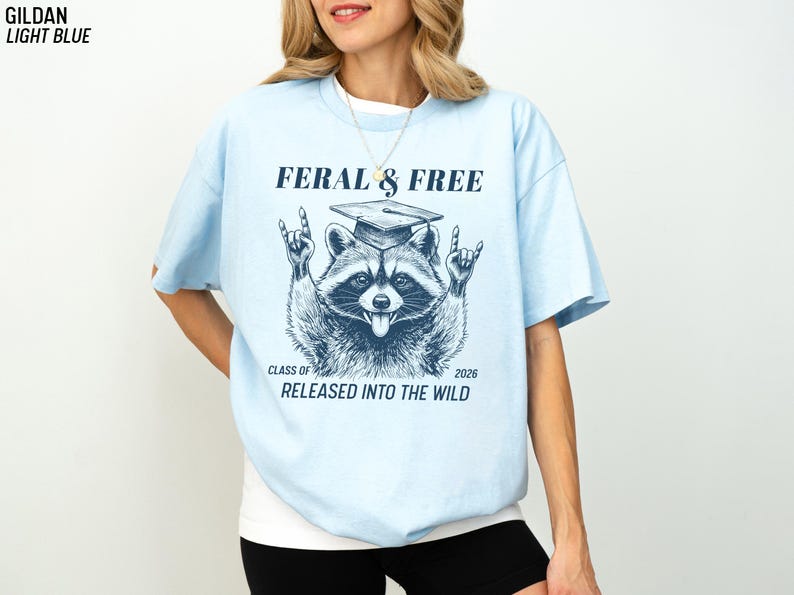 May include: Light blue t-shirt with a raccoon graphic wearing a graduation cap and the text "FERAL & FREE". The raccoon is making a rock and roll hand gesture. The text "CLASS OF 2026 RELEASED INTO THE WILD" is also printed on the shirt.