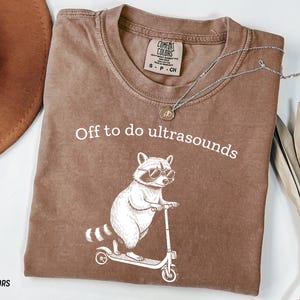 May include: Espresso-colored t-shirt with the text "Off to do ultrasounds" above a raccoon wearing sunglasses and riding a scooter. A silver necklace and a brown hat are also visible.