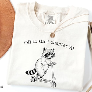 May include: Ivory Comfort Colors t-shirt with the text "Off to start chapter 70". The shirt features a black and white illustration of a raccoon wearing sunglasses and riding a scooter.