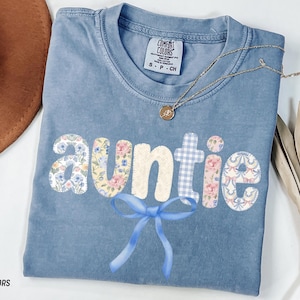 May include: A blue Comfort Colors t-shirt with the word "Auntie" in a floral and gingham pattern, with a blue bow. A gold necklace with a pendant is on the shirt. The shirt is folded.