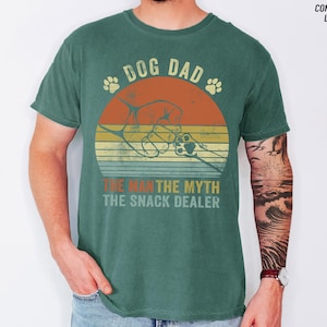 May include: A teal-colored t-shirt with a graphic design featuring a dog paw and the words "DOG DAD" above a retro sunset design. Below the sunset, the text reads "THE MAN THE MYTH THE SNACK DEALER."