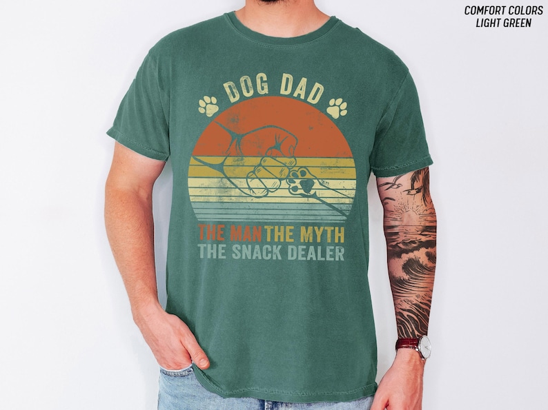May include: A teal-colored t-shirt with a graphic design featuring a dog paw and the words "DOG DAD" above a retro sunset design. Below the sunset, the text reads "THE MAN THE MYTH THE SNACK DEALER."