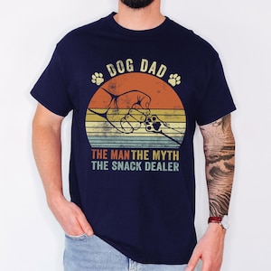 May include: Navy blue t-shirt with a retro design. The graphic features a dog paw print, a hand holding a dog's paw, and the text "DOG DAD THE MAN THE MYTH THE SNACK DEALER" in a vintage style. The shirt is made of a soft material.