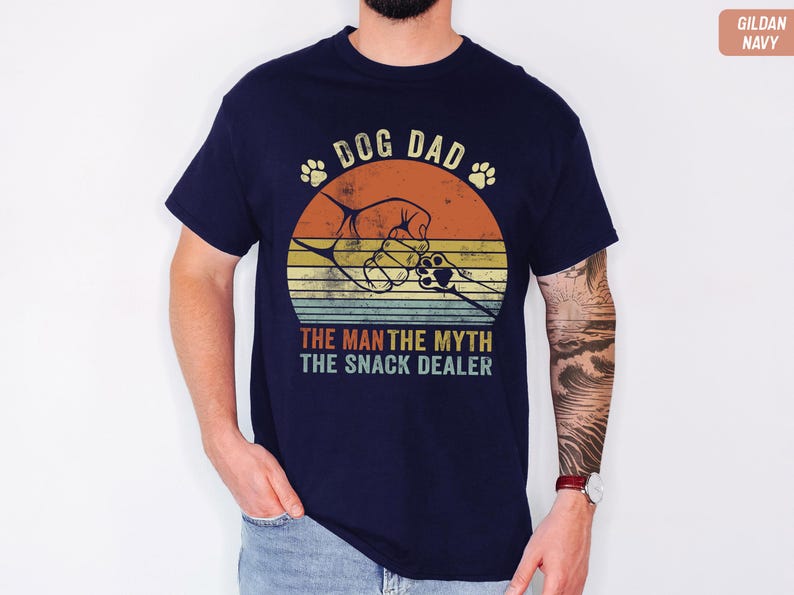 May include: Navy blue t-shirt with a retro design. The graphic features a dog paw print, a hand holding a dog's paw, and the text "DOG DAD THE MAN THE MYTH THE SNACK DEALER" in a vintage style. The shirt is made of a soft material.