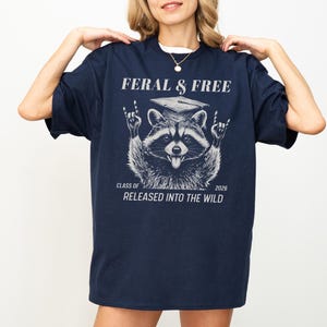 May include: Navy blue t-shirt with a raccoon graphic wearing a graduation cap and the text "FERAL & FREE". The raccoon is making a rock and roll hand gesture. The text "CLASS OF 2026 RELEASED INTO THE WILD" is also printed on the shirt.