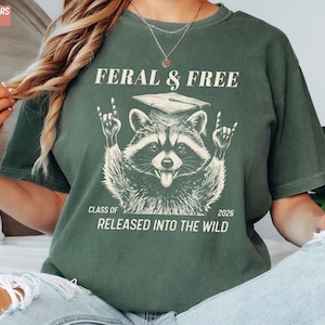 May include: Moss green t-shirt with a raccoon graphic wearing a graduation cap and the text "FERAL & FREE" above it. Below the raccoon is "CLASS OF 2026 RELEASED INTO THE WILD". The shirt is a comfort color.