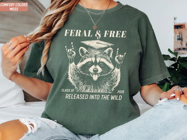May include: Moss green t-shirt with a raccoon graphic wearing a graduation cap and the text "FERAL & FREE" above it. Below the raccoon is "CLASS OF 2026 RELEASED INTO THE WILD". The shirt is a comfort color.
