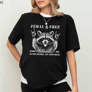 May include: Black Bella Canvas t-shirt with a raccoon graphic. The raccoon is giving the rock and roll hand sign. Text on the shirt reads "FERAL & FREE", "RETIRED 2026", and "NO MORE MEETINGS. JUST TRASH SNACKS".