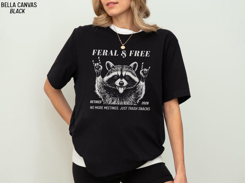May include: Black Bella Canvas t-shirt with a raccoon graphic. The raccoon is giving the rock and roll hand sign. Text on the shirt reads "FERAL & FREE", "RETIRED 2026", and "NO MORE MEETINGS. JUST TRASH SNACKS".