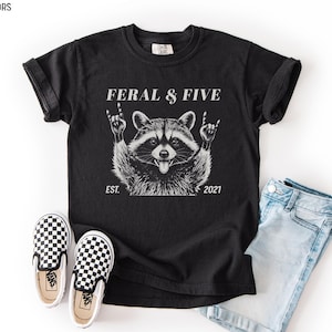 May include: Black t-shirt with a raccoon graphic, the text "FERAL & FIVE", and the year "2021". The raccoon is making a rock and roll hand gesture. Also pictured are checkered slip-on shoes and light blue denim shorts.