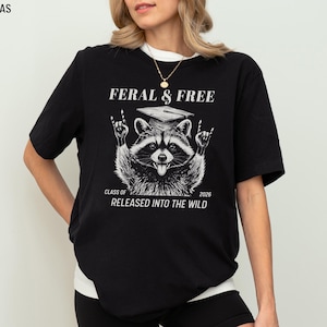 May include: Black t-shirt with a raccoon graphic wearing a graduation cap and making a rock-and-roll hand gesture. The text reads "FERAL & FREE" and "CLASS OF 2026 RELEASED INTO THE WILD."