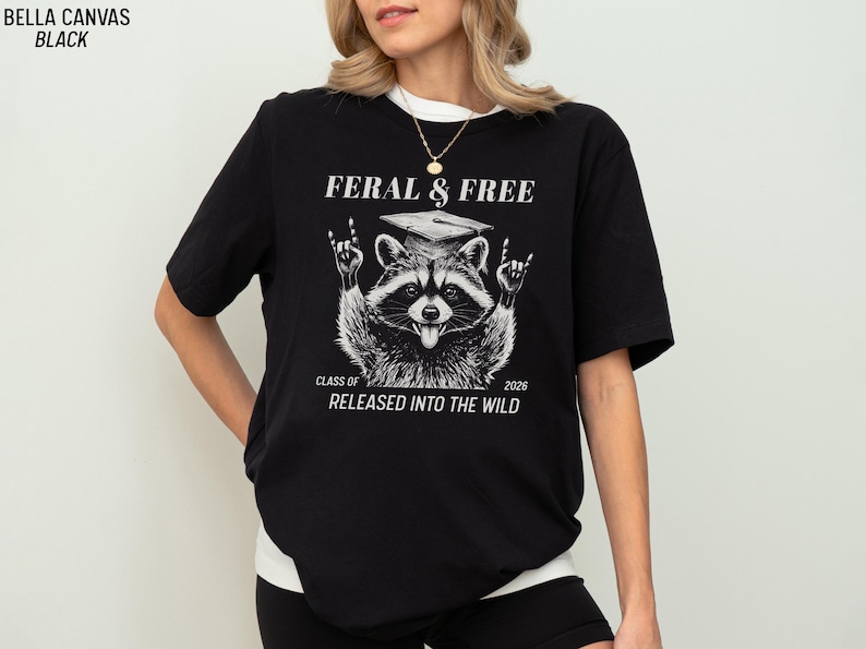 May include: Black t-shirt with a raccoon graphic wearing a graduation cap and making a rock-and-roll hand gesture. The text reads "FERAL & FREE" and "CLASS OF 2026 RELEASED INTO THE WILD."
