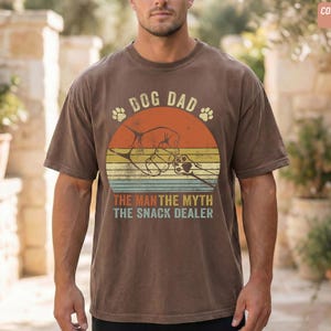 May include: Brown t-shirt with a graphic design featuring the text "DOG DAD" above a retro sunset design with paw prints and the text "THE MAN THE MYTH THE SNACK DEALER". The shirt is a comfort color in espresso.