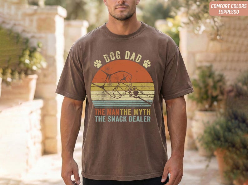May include: Brown t-shirt with a graphic design featuring the text "DOG DAD" above a retro sunset design with paw prints and the text "THE MAN THE MYTH THE SNACK DEALER". The shirt is a comfort color in espresso.