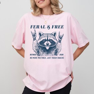 May include: A light pink t-shirt with the text "FERAL & FREE" above a raccoon graphic. Below the graphic are the words "RETIRED 2026" and "NO MORE MEETINGS. JUST TRASH SNACKS."