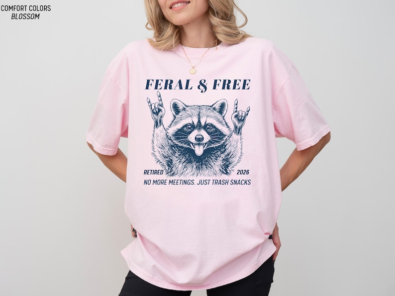 May include: A light pink t-shirt with the text "FERAL & FREE" above a raccoon graphic. Below the graphic are the words "RETIRED 2026" and "NO MORE MEETINGS. JUST TRASH SNACKS."