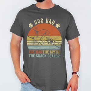 May include: Dark gray t-shirt with a retro design featuring a dog paw print, a hand, and the text "DOG DAD, THE MAN, THE MYTH, THE SNACK DEALER." The design is set against a sunset-style background with orange, yellow, and green stripes.