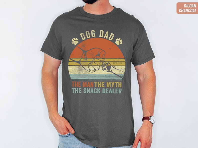 May include: Dark gray t-shirt with a retro design featuring a dog paw print, a hand, and the text "DOG DAD, THE MAN, THE MYTH, THE SNACK DEALER." The design is set against a sunset-style background with orange, yellow, and green stripes.