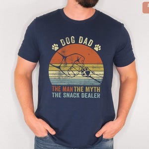 May include: Navy blue t-shirt with a retro graphic design. The design features a dog paw print, a fist bump, and the text "DOG DAD, THE MAN THE MYTH THE SNACK DEALER". The shirt is made of Bella Canvas material.