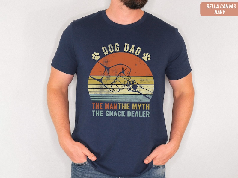 May include: Navy blue t-shirt with a retro graphic design. The design features a dog paw print, a fist bump, and the text "DOG DAD, THE MAN THE MYTH THE SNACK DEALER". The shirt is made of Bella Canvas material.