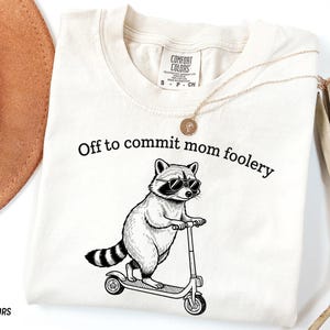 May include: Ivory-colored t-shirt with the phrase "Off to commit mom foolery" and a raccoon wearing sunglasses riding a scooter. The shirt is made of Comfort Colors material.