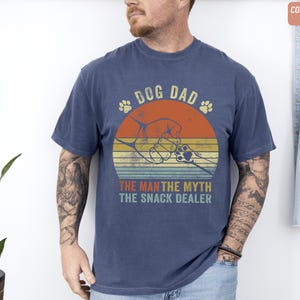 May include: Navy blue t-shirt with a retro design. The graphic features the text "DOG DAD" above a circular design with a dog paw and the text "THE MAN THE MYTH THE SNACK DEALER". The shirt is a comfort color.