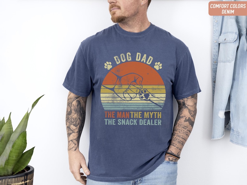 May include: Navy blue t-shirt with a retro design. The graphic features the text "DOG DAD" above a circular design with a dog paw and the text "THE MAN THE MYTH THE SNACK DEALER". The shirt is a comfort color.
