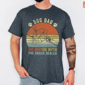 May include: Dark heather gray t-shirt with a retro design. The shirt features the text "DOG DAD" above a graphic of a hand clasping a paw, set against a sunset. Below, it reads "THE MAN THE MYTH THE SNACK DEALER."