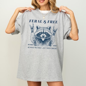 May include: A light gray t-shirt with a blue graphic of a raccoon making a rock and roll hand gesture. The text on the shirt reads "FERAL & FREE", "RETIRED 2026", and "NO MORE MEETINGS. JUST TRASH SNACKS."