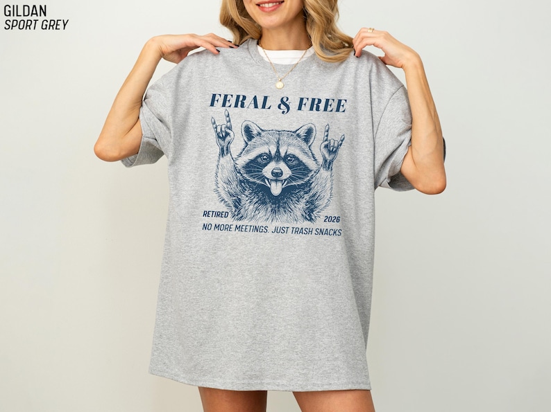 May include: A light gray t-shirt with a blue graphic of a raccoon making a rock and roll hand gesture. The text on the shirt reads "FERAL & FREE", "RETIRED 2026", and "NO MORE MEETINGS. JUST TRASH SNACKS."