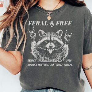 May include: Dark grey t-shirt with a raccoon graphic. The raccoon is making a rock and roll hand gesture. Text on the shirt reads "FERAL & FREE", "RETIRED 2026", and "NO MORE MEETINGS. JUST TRASH SNACKS."