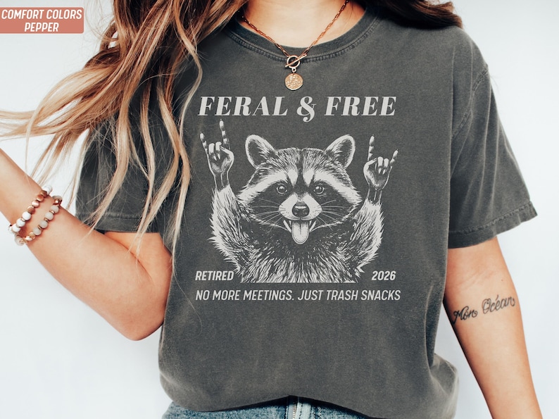 May include: Dark grey t-shirt with a raccoon graphic. The raccoon is making a rock and roll hand gesture. Text on the shirt reads "FERAL & FREE", "RETIRED 2026", and "NO MORE MEETINGS. JUST TRASH SNACKS."