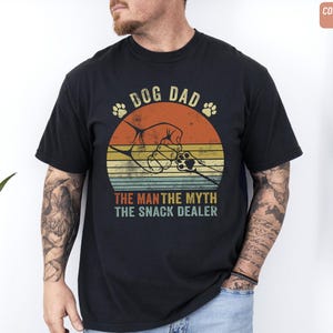 May include: Black t-shirt with a retro design featuring the text "DOG DAD" above a graphic of a dog paw and a dog's paw holding a hand. The text "THE MAN THE MYTH THE SNACK DEALER" is below the graphic. The shirt has a vintage, distressed look.