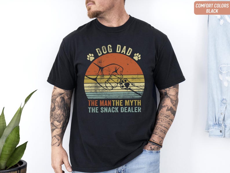 May include: Black t-shirt with a retro design featuring the text "DOG DAD" above a graphic of a dog paw and a dog's paw holding a hand. The text "THE MAN THE MYTH THE SNACK DEALER" is below the graphic. The shirt has a vintage, distressed look.